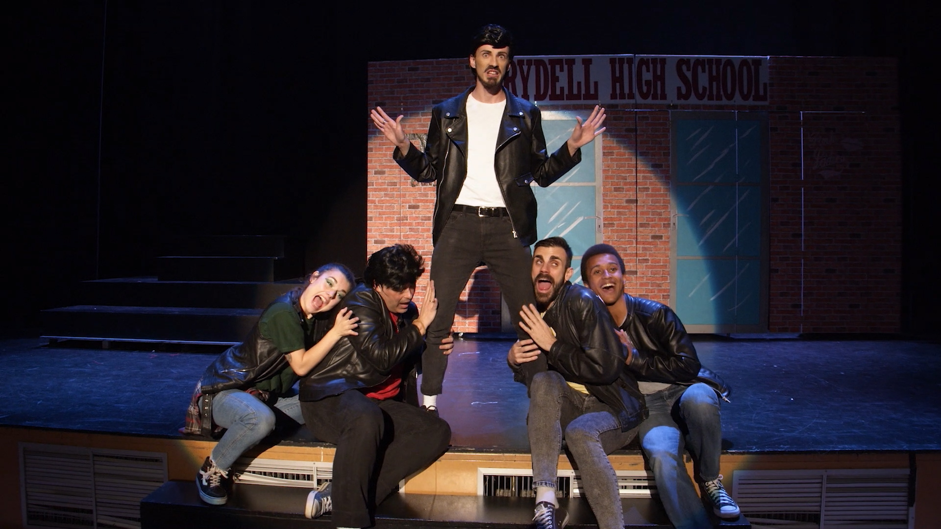 un grupo de jóvenes haciendo teatro musical en un escenario que imita una patio de colegio iluminados por un foco