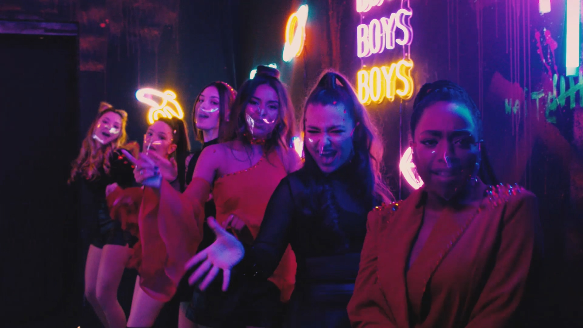 seis mujeres jóvenes con mascarillas transparentes en un espacio con letreros de neón cantando hacia la cámara