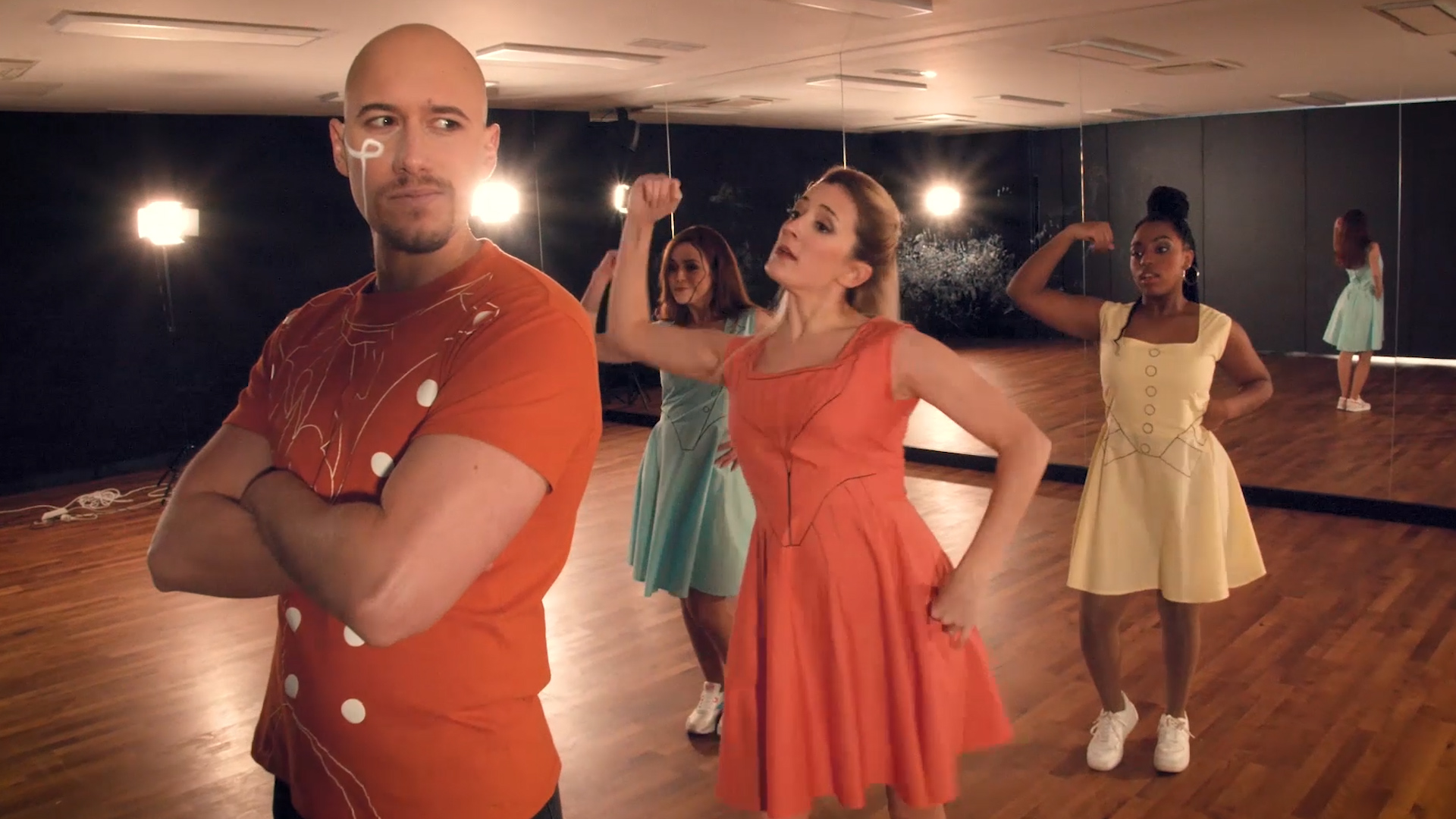 tres mujeres bailan y cantan con un hombre con los brazos cruzados mirando de reojo en primer plano
